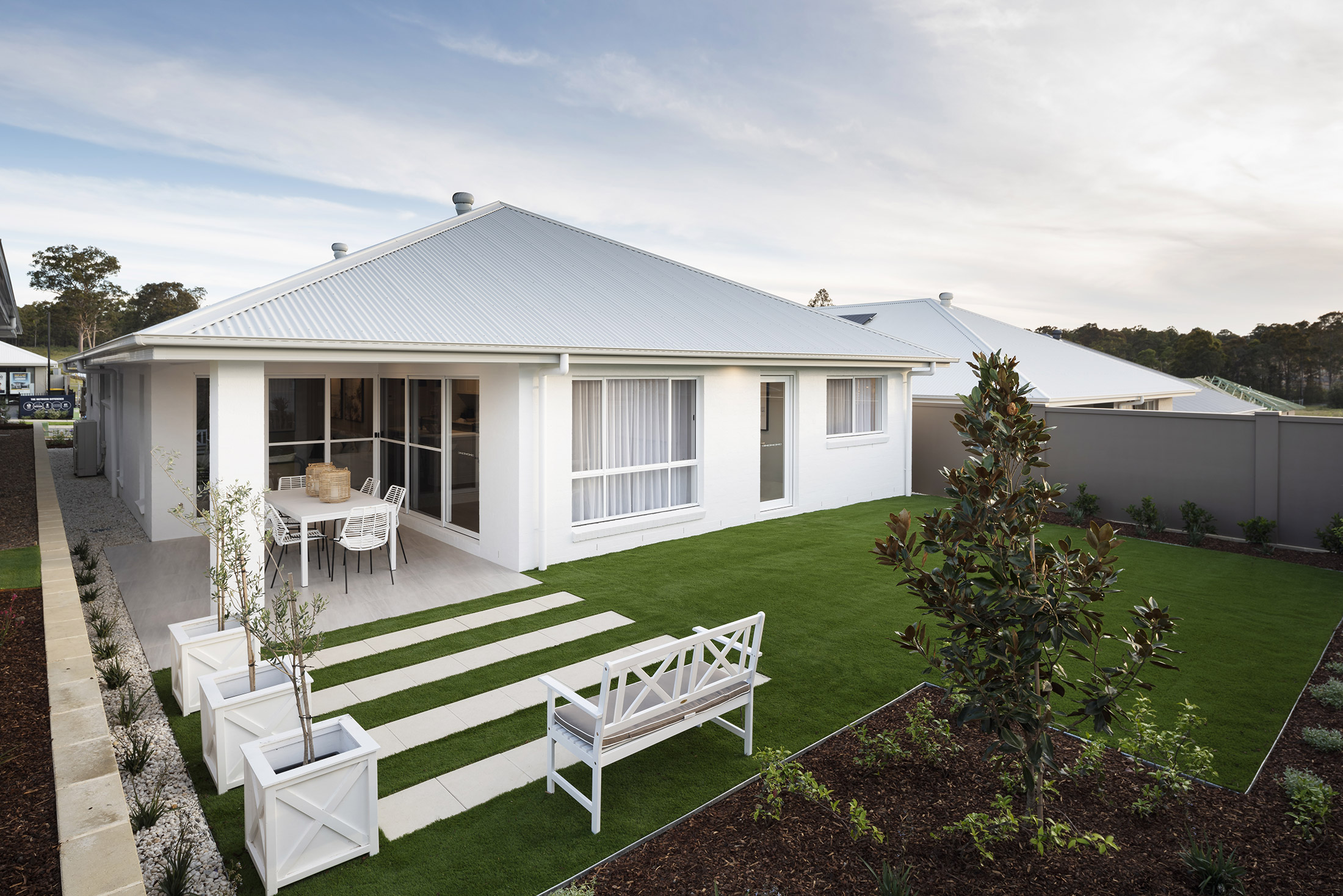Backyard of the Avoca display home featuring grassed area, wooden bench overlooking stoned pavers and facing a larger paved dining area
